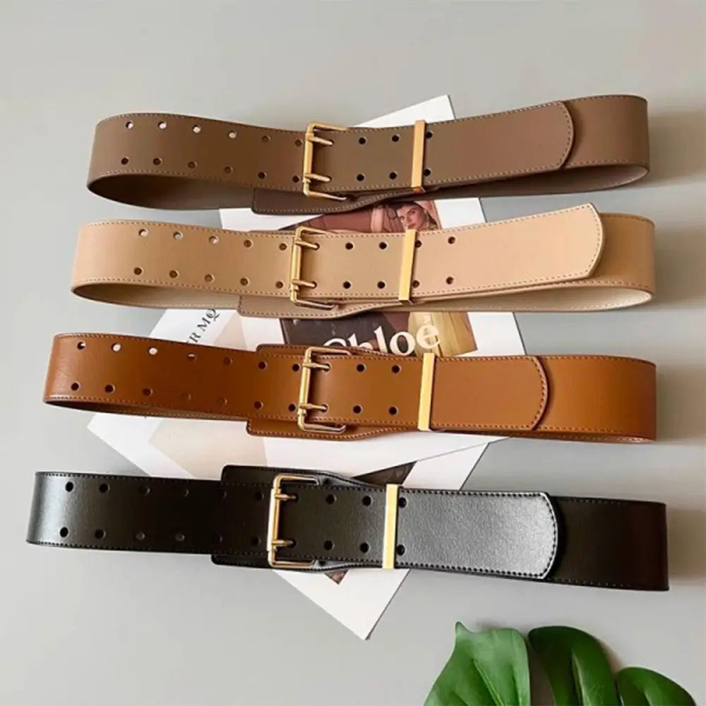 Four wide leather belts with gold buckles displayed with a beige turtleneck sweater and bag